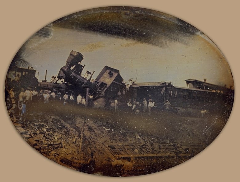 Old photo of a crowd gathered around a train that has gone off its rails.