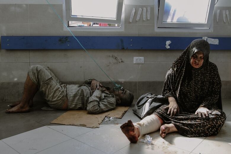 A woman in a headscarf is shown sitting on the ground with a wrap around her right leg and damage to her face. Beside her a man lies on the ground - he is covered in dirt and has an oxygen mask attached to his face.