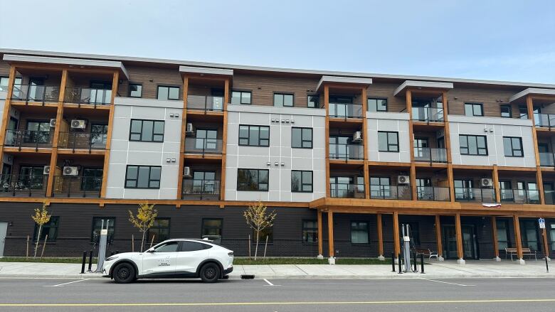 A four-story apartment building sits on a concrete street with a white SUV parked at an electrical charging station in front.