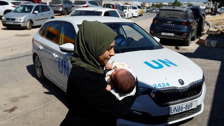 A woman carries a small baby in her arms as she walks through a parking lot. A vehicle marked for the United Nations is in the background.