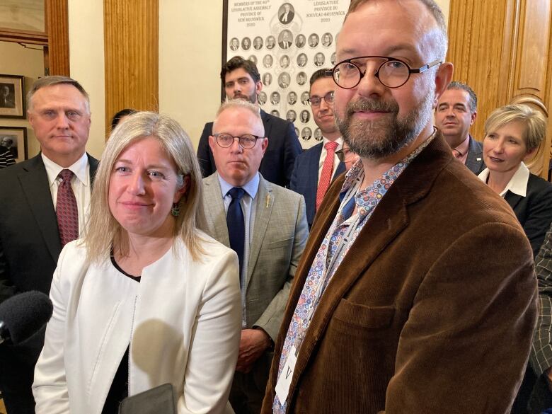 A woman in a white blazer and a tall man with glasses stand in front of a microphone in a crowded hallway.