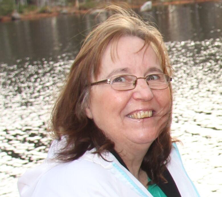 A woman wearing glasses smiles and looks toward the camera. Water and trees can be seen in the background.