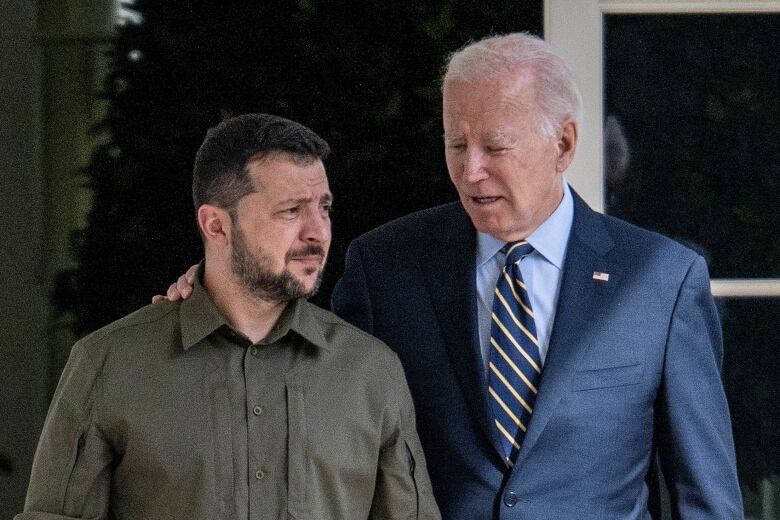 Grey haired man in blue suit standing with arm around the shoulder of dark haired man in khaki military wear