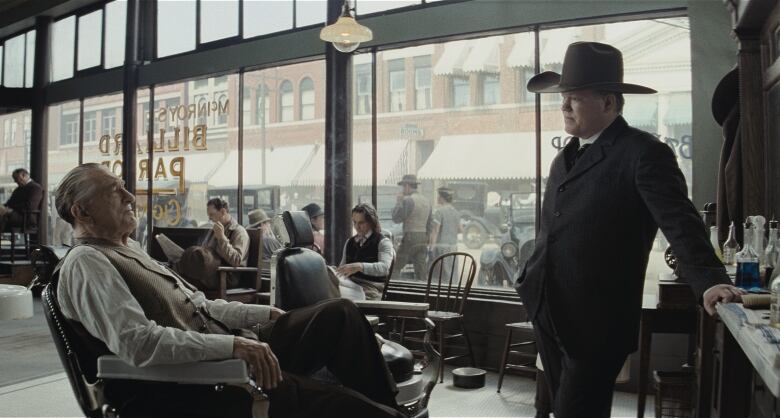 An FBI agent wearing a cowboy hat questions an old man sitting in a barbershop chair. 