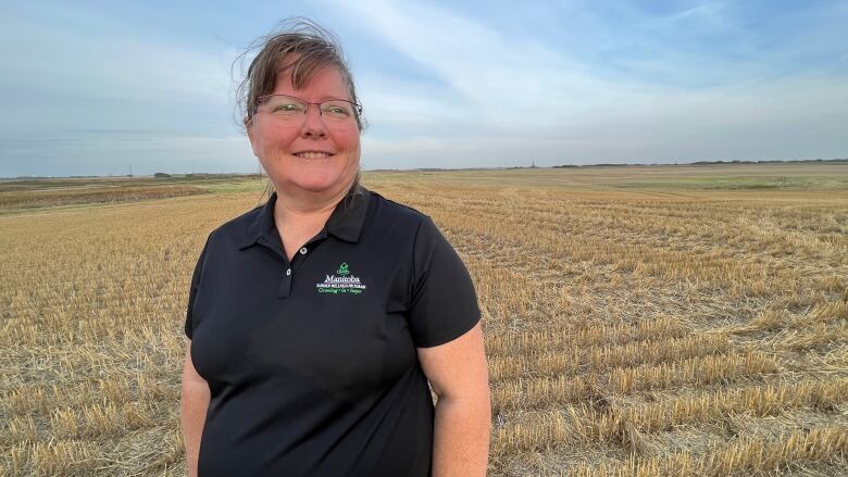 A woman with glasses, wearing a black shirt stands in a field of stubble in front of a mainly clear blue sky while smiling and looking into the distance.