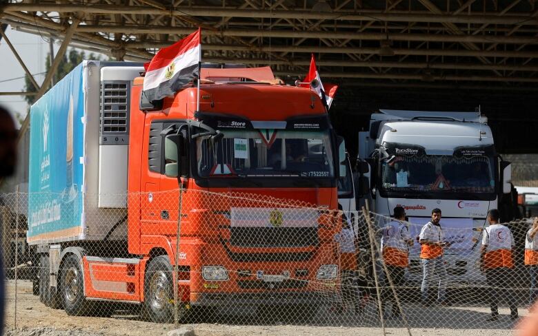 A transport truck with a bright orange cab, with a red, white and black flag affixed on the front and two more on the roof, is parked beside another white truck with people standing in front.