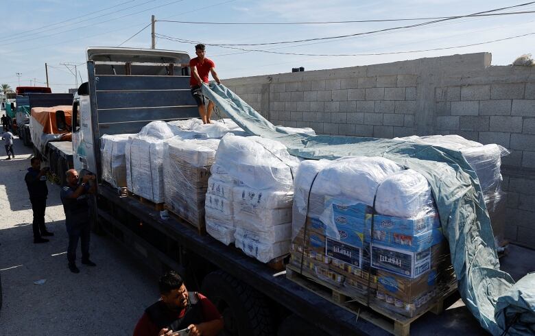 A man pulls a tarp over stacks of boxes loaded on the back of a flatbed truck. 