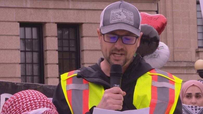 A man wearing a high-visibility vest, glasses and a hat speaks into a microphone.