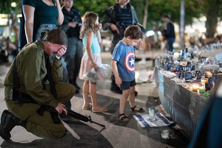 A young woman in green army fatigues and carrying an automatic rifle cries as she kneels before a candlelit makeshift memorial.