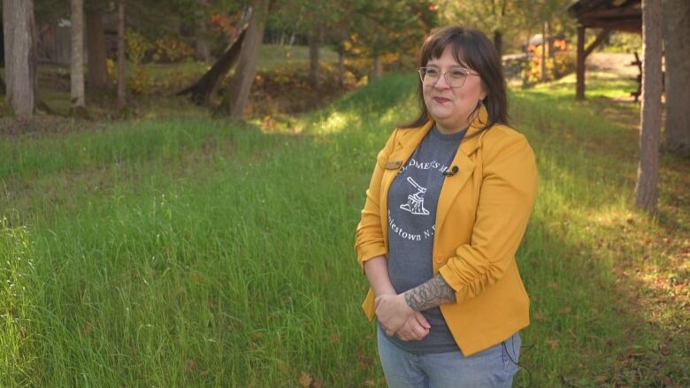 A smiling woman in a yellow blazer with her arms crossed in front over her stomach. She is stood next to a berm which has long grass.