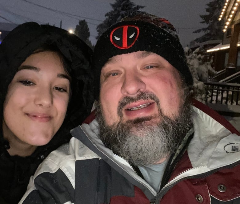 A teenage girl and her father smile for a selfie. They are dressed warmly, standing outside in winter with snow-covered trees in the background.