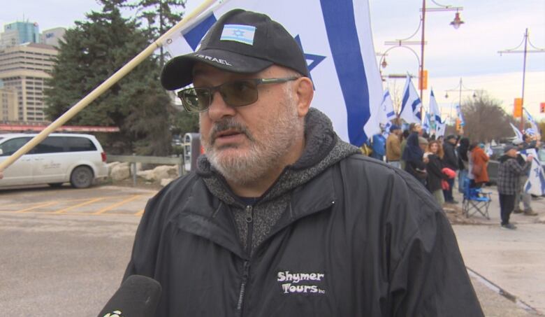 A man in a black ball cap with the Israel flag on it, wears a black jacket and glasses. He has a white beard.