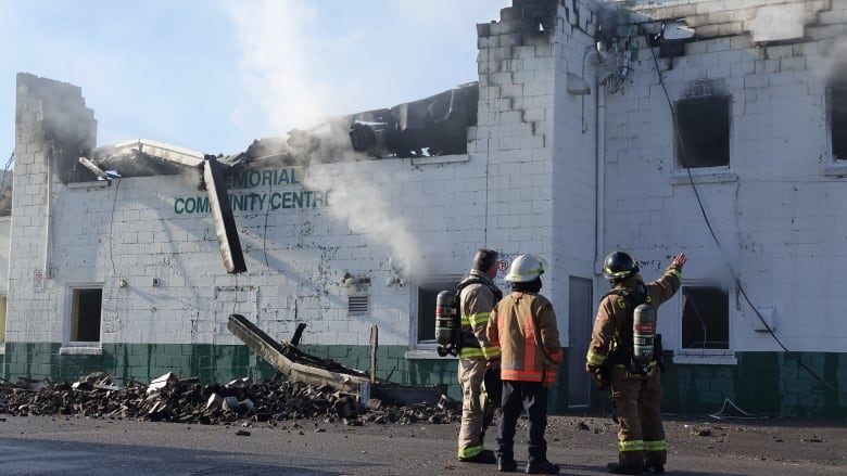 Three firefighters in full gear stand in front of a white and green brick building that's partially collapsed. Smoke is in the air and rubble is one the ground.