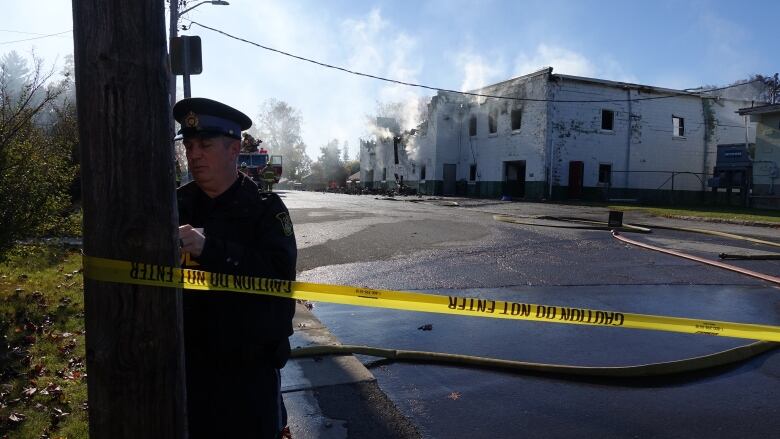 A police officer ties yellow caution tape to a light pole. In the background a white and green brick building that's partially collapsed can be seen smoking.