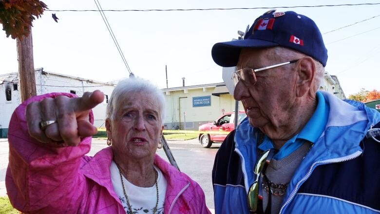 A small elderly woman in a pink coat stands next to an older man in a blue jacket. She's pointing toward the camera with a distressed look on her face.