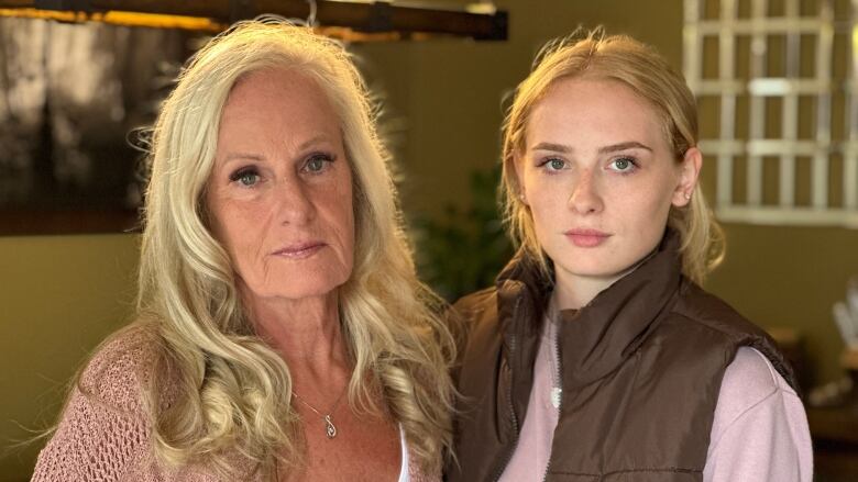 An older woman with blond hair stands beside a young woman. Both wear neutral expressions.