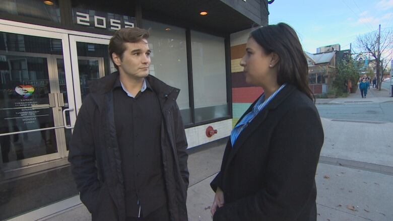 A man and a woman stand on a sidewalk facing each other wearing dark jackets. 