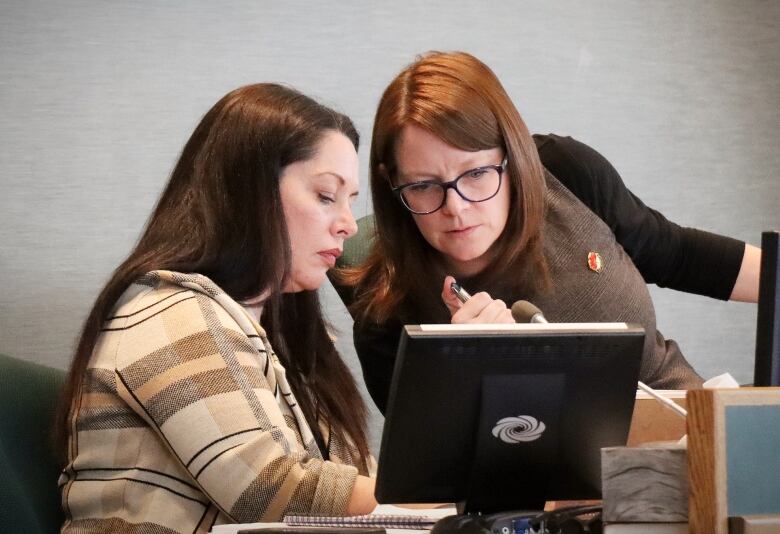 Two women, one with red hair and glasses and one with brown hair, put their heads together looking at something behind a computer screen.