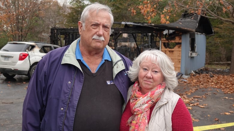 A tall man with white hair and a moustache stands next to a small woman in a white vest and orange sweater. Behind them is a burned down garage and a vehicle with scorch marks on it.