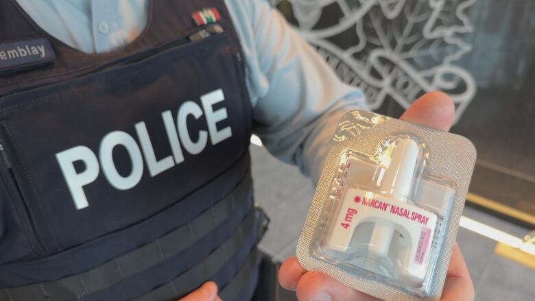 A police officer is shown wearing a blue vest and holding a narcan naloxone nasal spray in his hand.