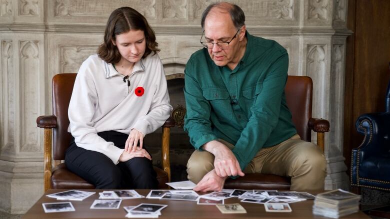 a women in a white sweater wearing a poppy sits with a man wearing a dark green shirt while sorting black and white photos 