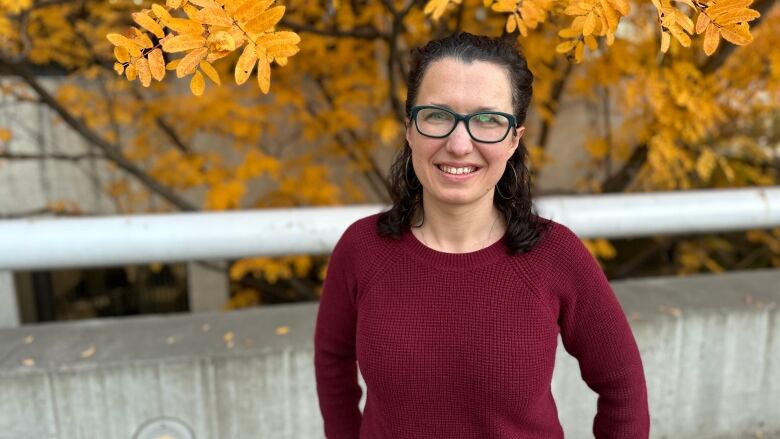 A brunette woman in a burgundy shirt stands in front of a tree with autumn colours. 