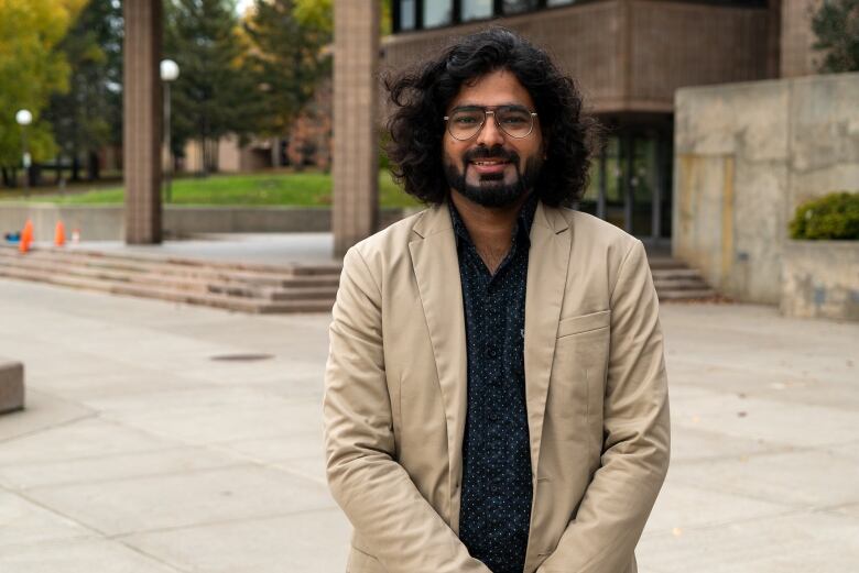 A man with dark hair and glasses wearing a sport coat stands in front of a campus building. 