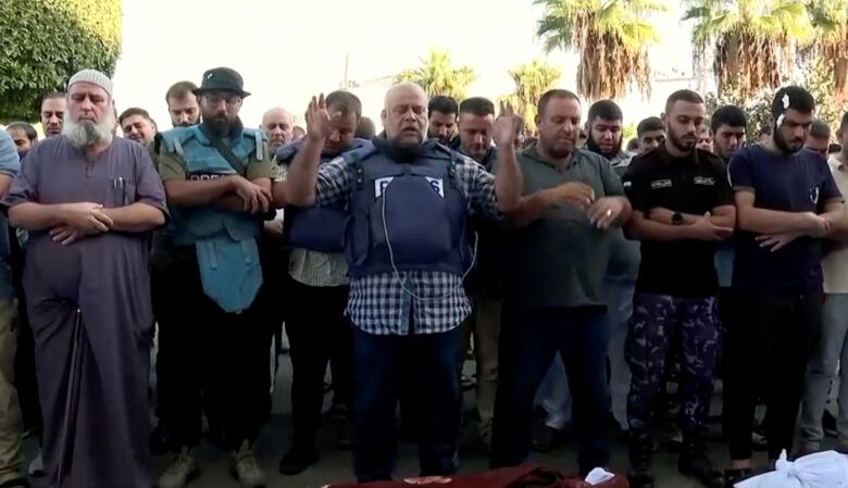 Several men are shown with solemn looks on their face. A man in the middle, wearing a blue vests, raises his arms in a prayer-like gesture.