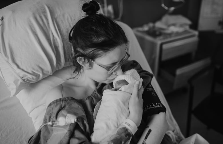 A woman sits in a hospital bed holding a baby and kissing its forehead.