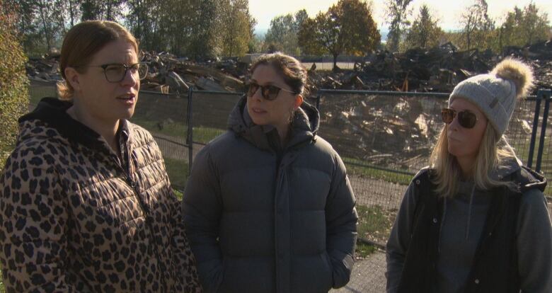 Three women in warm clothing stand in front of a fence cordoning off rubble from a fire.