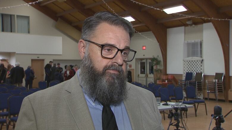 A man with glasses, suit and full beard stands during an interview.