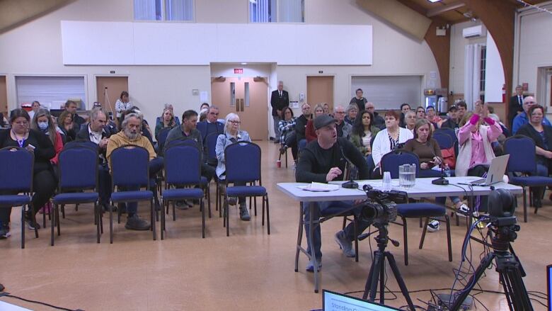 A man with a flat-brim hat and black shirt sits at a table speaking with MLAs.