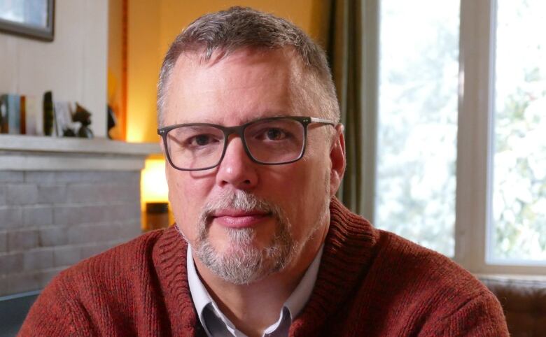 A man wearing glasses, a white dress shirt and rust-coloured sweater sits in an indoor room, with a fireplace and window seen behind him.