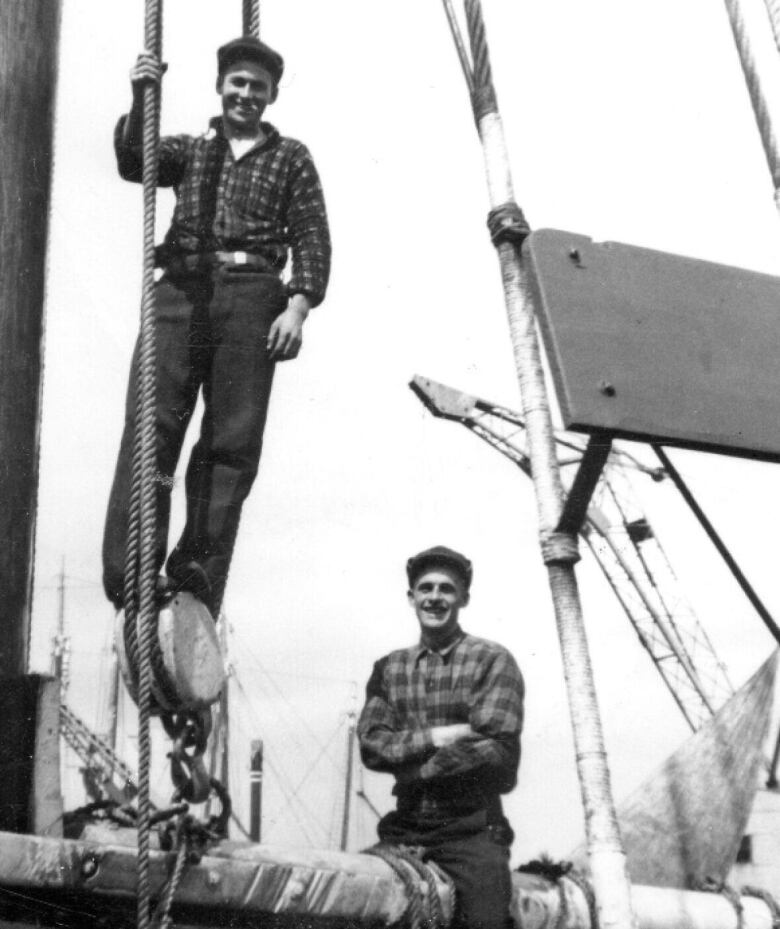 An archival photograph shows a young man standing along a wooden mast. 