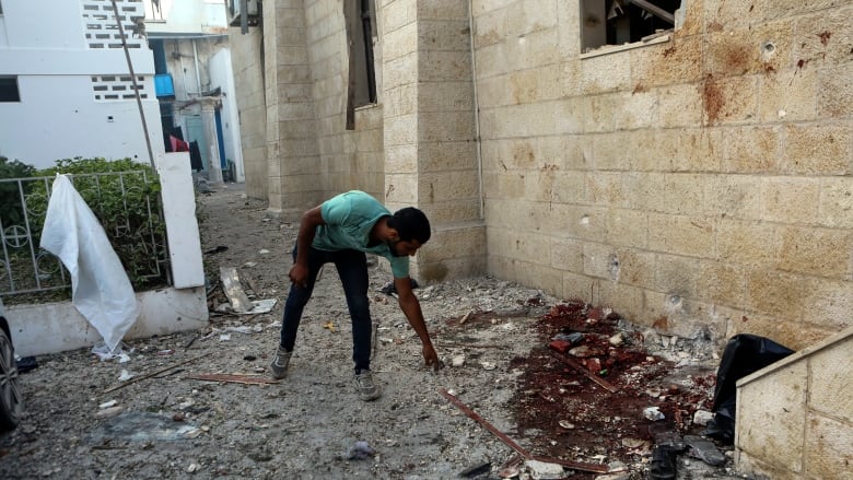 A man points on the ground filled with debris and a large stain next to a building wall.