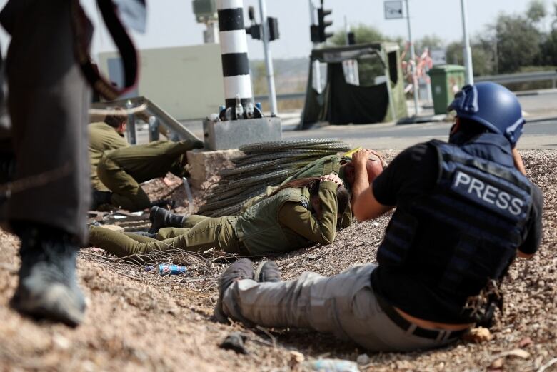 A photographer wearing a 'PRESS' vest snaps images of soldiers taking cover on the ground.