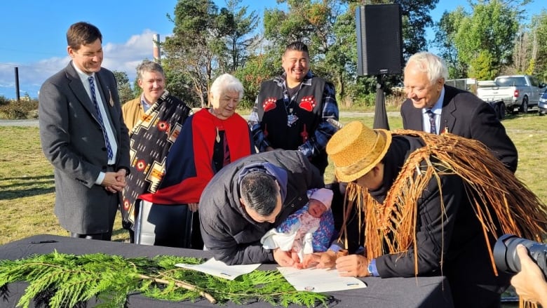 A group of smiling adults watch as a father holds his infant's inked foot to a document so she can sign it. 