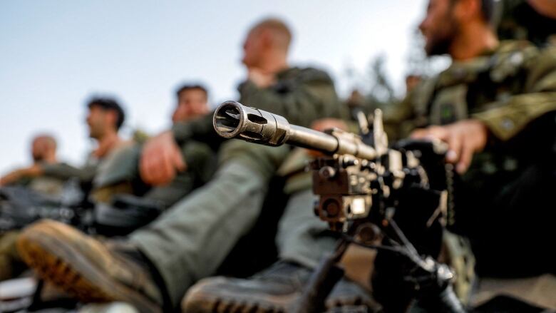 A machine gun is seen in focus while out-of-focus soldiers are seated behind the weapon.