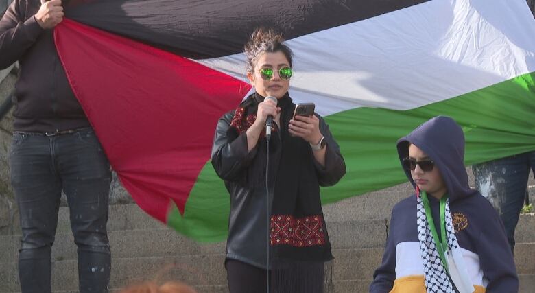 A woman with dark hair and sunglasses holds a microphone and a smartphone standing in front of a Palestinian flag. 