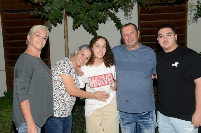 Five people are seen standing in a line for a photo. A young woman with curly hair wearing a white T-shirt is in the middle.