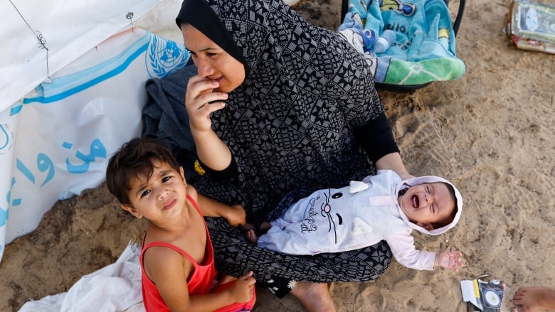 A woman pictured from above sitting the sand beneath a tent with a small child next to her and a crying baby in her arms. 