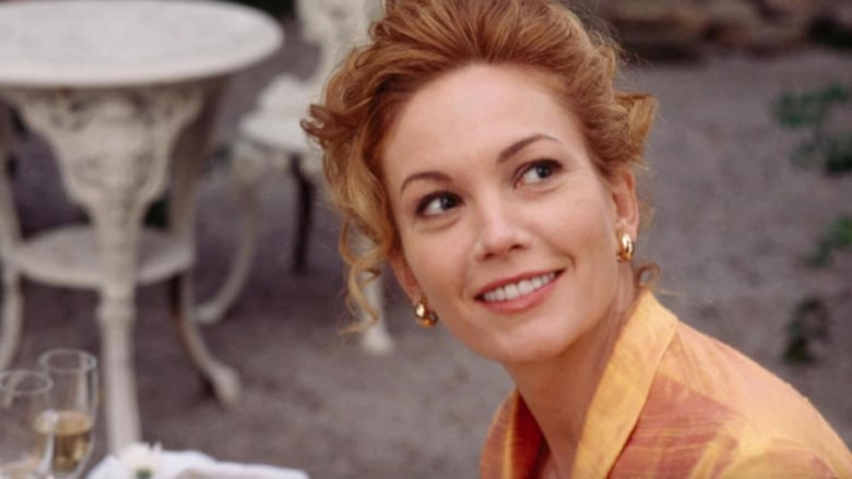 Closeup on Diane Lane in the movie Under the Tuscan Sun. She's wearing an orange shirt, sitting outside at a table with wine glasses on it and smiling into the distance. 