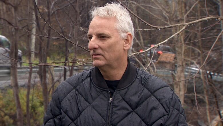 A white man with a black jacket stands outside near a road, with trees behind him