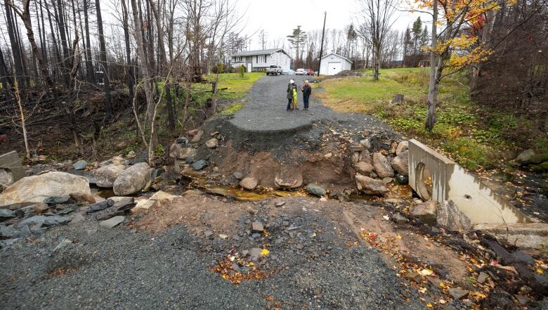 A large crack in the earth of a driveway exposes the layers of gravel and dirt above a brook. Three people stand a few feet up the driveway away from the gap, closer to a bungalow in the distance