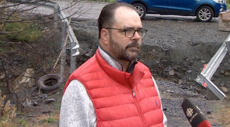 A white man with glasses and a beard wears a grey sweater under a red puffy vest. He is standing outside by a road, and the gap in the driveway is behind him