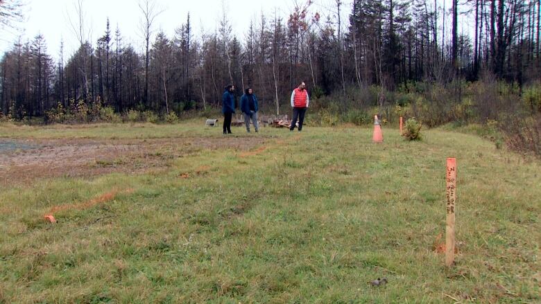 A man in a red vest stands alongside two other men in blue jackets and sweaters on a grassy lawn with orange spraypaint in the grass and wooden stakes