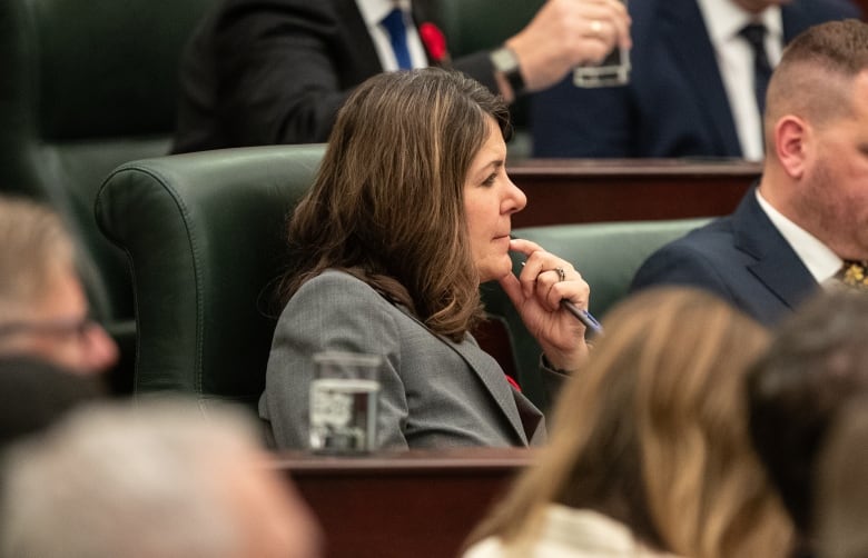 A woman holds a pen and touches her face with the same hand while sitting in a legislature chamber.