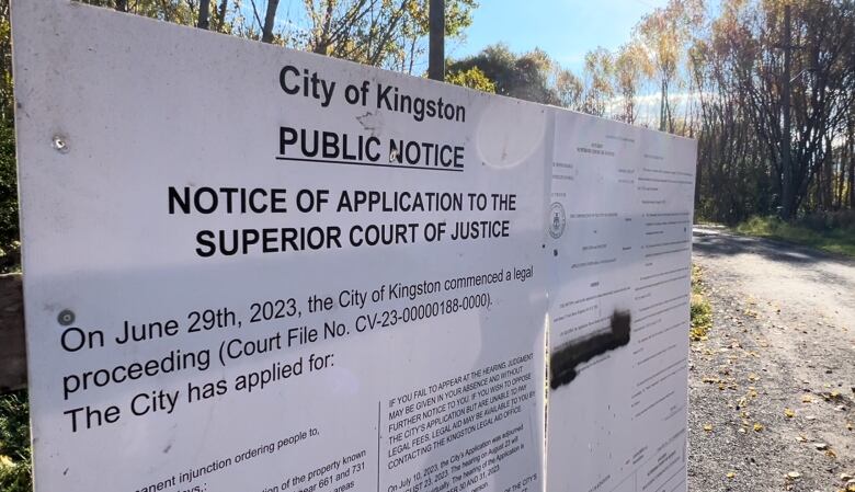 A legal document is printed out on two large, white boards. It explains Kingston is seeking an injunction to clear the park. It's a sunny, fall day and a gravel path and trees with yellow leaves can be seen in the background.