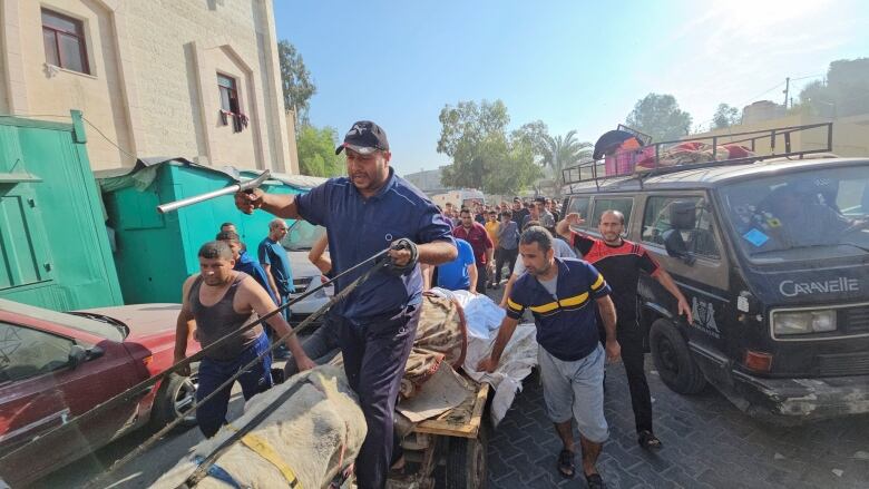 A group of people following a donkey-drawn cart, next to a car. 