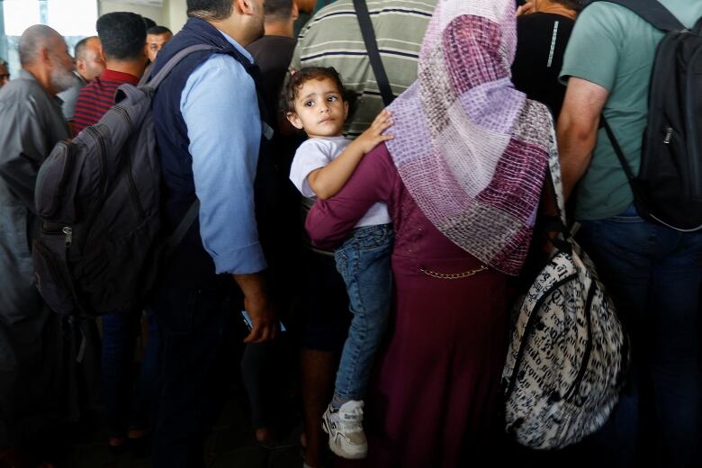 A woman holds a toddler on her hip as they wait in a crowded room at a border crossing.